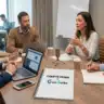 Une réunion de travail professionnelle se déroule autour d'une table en bois dans une salle lumineuse. Cinq collaborateurs échangent activement autour d'un microphone de table, de ordinateurs portables et de tasses à café. Au premier plan, un bloc-notes à spirales blanc est posé sur la table, affichant en son centre la mention « COMPTE RENDU by Gaia Scribe » avec le logo officiel de la marque. En arrière-plan, une autre personne travaille sur un ordinateur et un tableau blanc affiche des diagrammes flous.