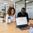 Une photographie grand angle d'un bureau moderne et lumineux en espace ouvert avec trois employés de bureau, deux assis et un troisième de profil, se faisant face. Une femme avec une coiffure bouclée présente son smartphone qui affiche un code de sécurité sur l'écran pour l'authentification multifacteur, et un homme avec des lunettes regarde un ordinateur portable qui montre également une demande d'authentification multifacteur. Un homme sur la droite regarde la femme. Généré par Gemini